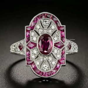 May include: An Art Deco-style ring with a central oval ruby surrounded by smaller rubies and diamonds. The ring has a silver band and a geometric design, showcasing a blend of vintage and modern aesthetics. The ring's design is symmetrical and elegant.