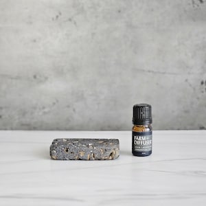 May include: A small black bottle with a black label that says "Farm Diffuser" and "Green Lavender Essential Oil" on a white surface next to a dark gray rectangular diffuser stone.