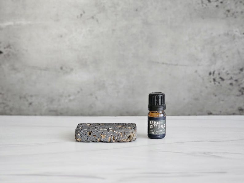 May include: A small black bottle with a black label that says "Farm Diffuser" and "Green Lavender Essential Oil" on a white surface next to a dark gray rectangular diffuser stone.