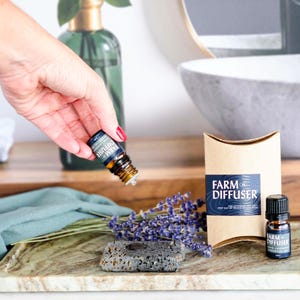 May include: A brown paper box with the text "FARM DIFFUSER" on it. The box is next to a small bottle of essential oil with a black cap and the text "FARM DIFFUSER 100% Lavender Essential Oil" on the label. A hand is holding a similar bottle of essential oil with the text "FARM DIFFUSER" on the label. The bottles are on a marble tray with a small gray stone diffuser and lavender flowers.