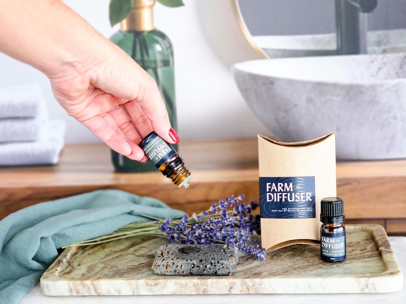 May include: A brown paper box with the text "FARM DIFFUSER" on it. The box is next to a small bottle of essential oil with a black cap and the text "FARM DIFFUSER 100% Lavender Essential Oil" on the label. A hand is holding a similar bottle of essential oil with the text "FARM DIFFUSER" on the label. The bottles are on a marble tray with a small gray stone diffuser and lavender flowers.