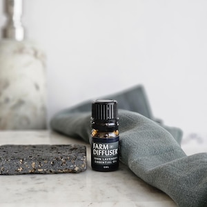 May include: A small, dark brown glass bottle with a black cap and label that reads "FARM DIFFUSER 100% LAVENDER ESSENTIAL OIL 10 ML" sitting on a gray cloth and a black stone coaster.