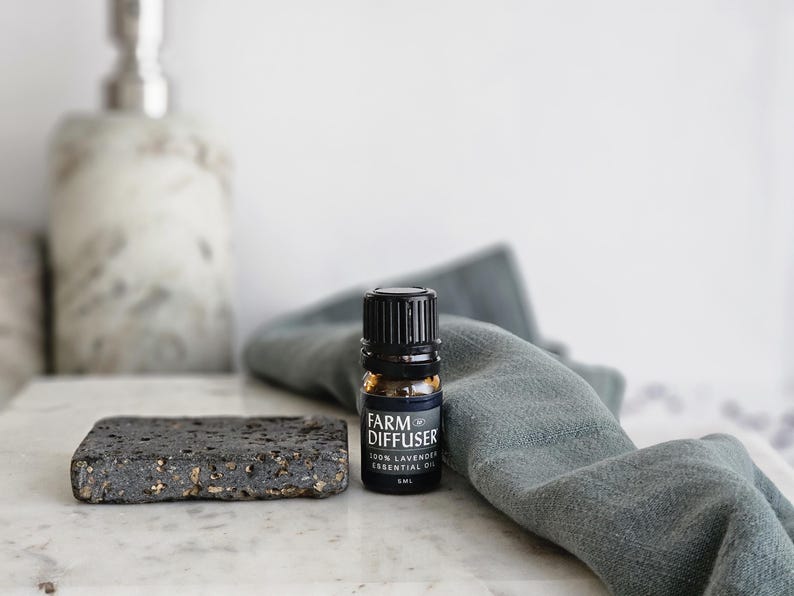 May include: A small, dark brown glass bottle with a black cap and label that reads "FARM DIFFUSER 100% LAVENDER ESSENTIAL OIL 10 ML" sitting on a gray cloth and a black stone coaster.