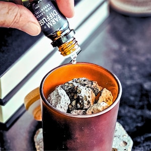 May include: A brown ceramic diffuser with a dark orange rim is filled with gray and brown lava rocks. A hand is pouring essential oil from a small glass bottle labeled "FARM DIFFUSE 100% LAVENDER ESSENTIAL OIL 5ML" into the diffuser.