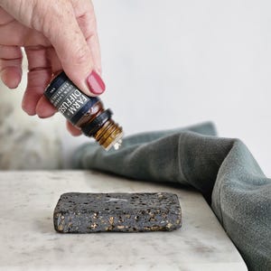 May include: A hand holding a small brown glass bottle with a black label that reads "FARM DIFFUSER LEMON LAVENDER ESSENTIAL OIL". The bottle is being poured onto a dark gray, textured stone diffuser.