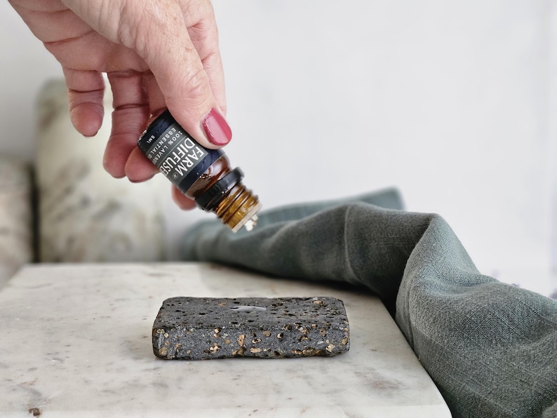 May include: A hand holding a small brown glass bottle with a black label that reads "FARM DIFFUSER LEMON LAVENDER ESSENTIAL OIL". The bottle is being poured onto a dark gray, textured stone diffuser.