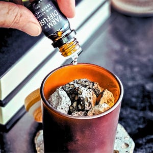 May include: A dark brown glass container with an orange interior is filled with various rocks. A small bottle labeled "FARM DIFFUSE" is pouring liquid into the container. The image suggests aromatherapy or essential oil use.