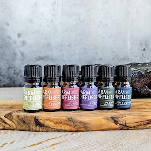 May include: Six small essential oil bottles with black caps are arranged on a wooden board. Each bottle has a label with the words "Farm Diffuser" and a unique scent name like "Bloom" or "Ignite". The background is a textured gray surface.