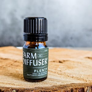 May include: A small, amber-colored glass bottle with a black cap. The label reads "FARM DIFFUSER" and "PLENTY" with the tagline "CALL IN WARMTH CULTIVATE ABUNDANCE." The bottle is 5ml and sits on a wooden surface.