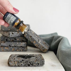 May include: A hand holding a small brown glass bottle with a black label that reads "AROMA DIFFUSER ESSENTIAL OIL 100% PURE", dropping oil onto a stack of black lava rock coasters.
