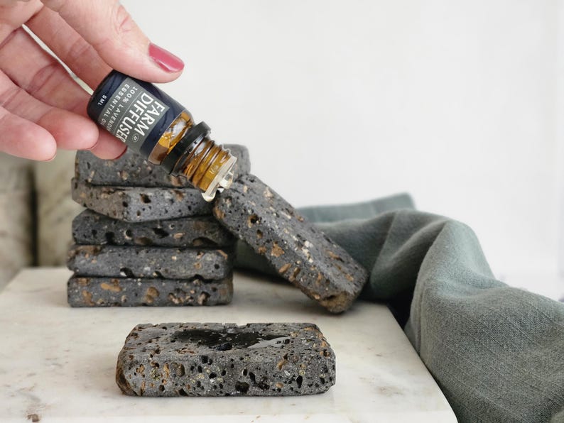 May include: A hand holding a small brown glass bottle with a black label that reads "AROMA DIFFUSER ESSENTIAL OIL 100% PURE", dropping oil onto a stack of black lava rock coasters.