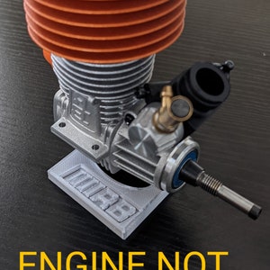 May include: A model engine with an orange cooling head and silver body, mounted on a gray base. The text "ENGINE NOT INCLUDED" is displayed in yellow at the bottom of the image. The engine is designed for model vehicles.