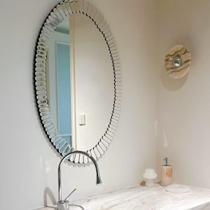 May include: An oval wall mirror featuring a decorative frame composed of numerous small, clear glass segments arranged in a radial pattern. The mirror reflects a doorway and interior space, positioned above a chrome faucet and marble countertop.
