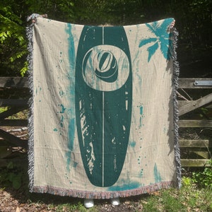 Pode incluir: Uma manta de tecido tecido de cor creme com um design de prancha de surf azul-esverdeado. A prancha de surf tem um círculo branco com um redemoinho azul-esverdeado dentro. A manta tem franjas ao redor das bordas.