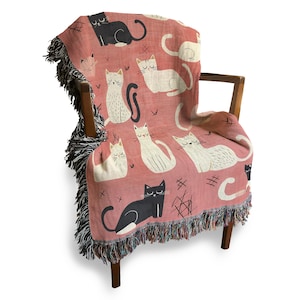May include: A pink woven throw blanket with a fringe border features a repeating pattern of black and white cats. The blanket is draped over a wooden chair.
