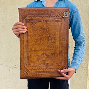 Puede incluir: Un libro grande, rectangular, encuadernado en cuero marrón con el título "Book of Shadows" en relieve en la portada. La portada presenta diseños de pentagrama y media luna. El libro tiene un cierre de metal.