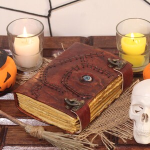 Op de afbeelding: Een bruin lederen gebonden boek met een gestikt oog op de omslag, omringd door Halloween-decoraties zoals een pompoen met een heksenhoed, een schedel en twee brandende kaarsen.