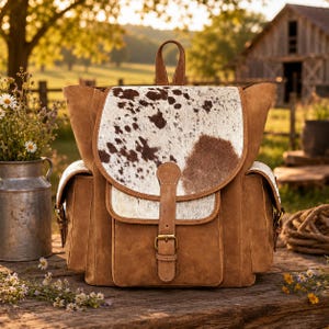 May include: A brown suede backpack featuring a cowhide flap with brown and white spots. The bag includes a top handle, two side pockets, and a central vertical strap with a gold-toned metal buckle closure. The design is structured with visible stitching.