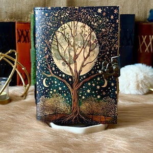 May include: A decorative journal with a celestial design featuring a large tree, moon, and stars. The cover is dark blue with a bronze clasp. The journal is resting on a white tray.