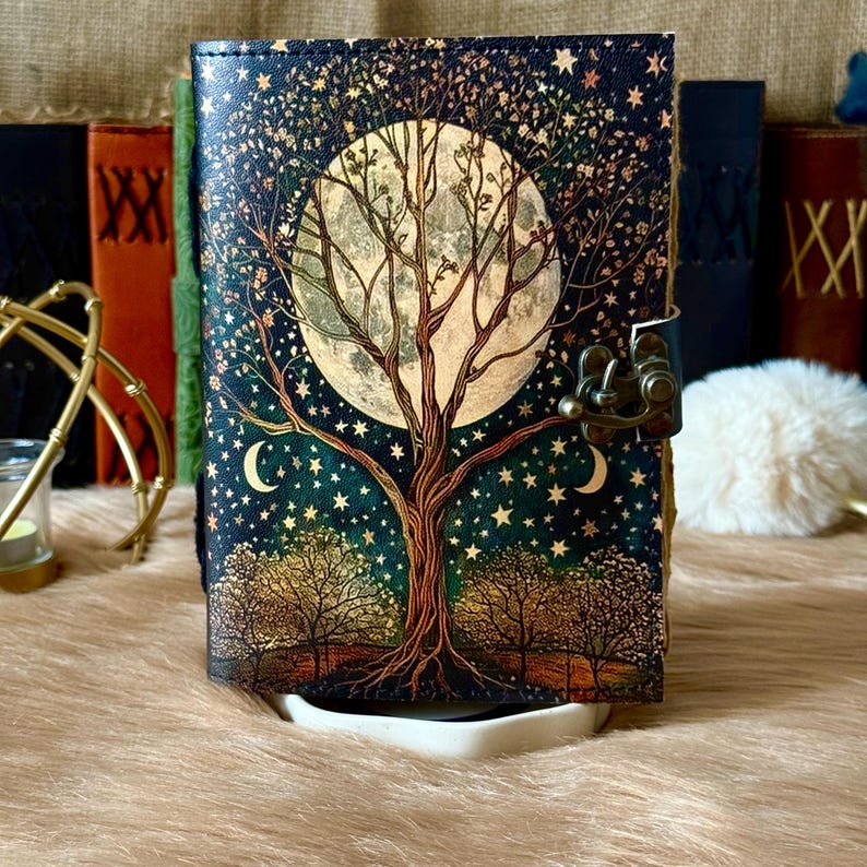 May include: A decorative journal with a celestial design featuring a large tree, moon, and stars. The cover is dark blue with a bronze clasp. The journal is resting on a white tray.