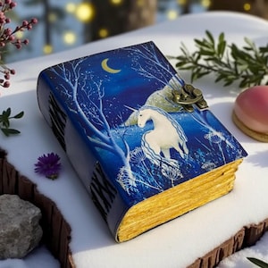 Può includere: Un diario rilegato in pelle blu con un'illustrazione di un unicorno sotto una luna crescente. Il diario ha bordi dorati e una chiusura in metallo. L'opera d'arte raffigura una scena notturna della foresta.