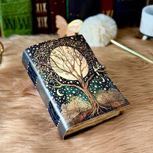May include: A leather journal with a celestial design. The cover shows a full moon, a tree, stars, and crescent moons on a dark blue background. The journal has a metal clasp and a dark blue spine.