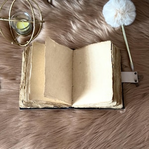 May include: An open, antique-style journal with aged, cream-coloured pages. The journal has a dark blue cover and a leather strap closure. A gold-coloured, spherical candle holder and a decorative pen with a white pom-pom are in the background.