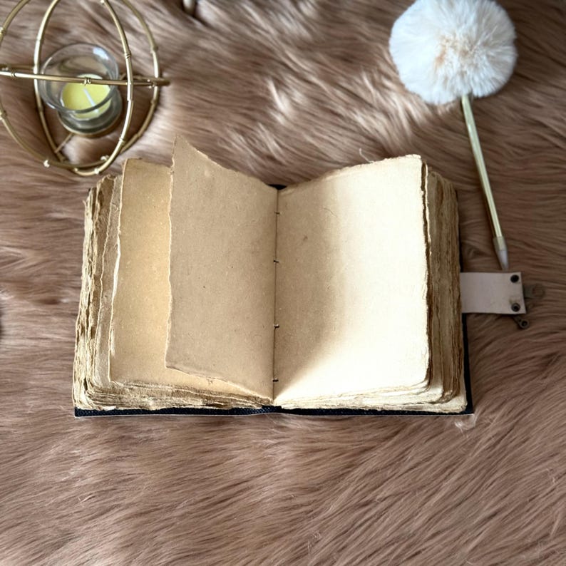 May include: An open, antique-style journal with aged, cream-coloured pages. The journal has a dark blue cover and a leather strap closure. A gold-coloured, spherical candle holder and a decorative pen with a white pom-pom are in the background.