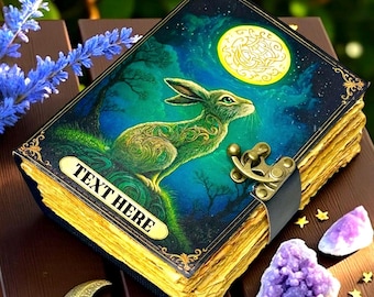 May include: A fantasy-themed journal with a green rabbit illustration under a full moon. The cover features the text "TEXT HERE" on a gold label. The journal has a clasp closure and aged-looking pages. Amethyst crystals and gold stars are scattered around the journal.