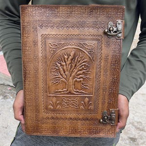 May include: A brown leather journal with a tree of life design embossed on the cover. The journal has a decorative border and a metal clasp closure. The leather has a natural, aged appearance, and the journal is held open.