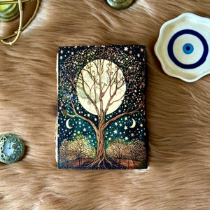 May include: A decorative journal with a celestial design featuring a full moon, a tree, stars, and crescent moons. The cover has a dark blue background with gold accents. The journal is lying on a brown, furry surface.