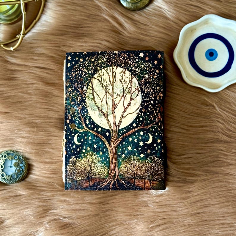 May include: A decorative journal with a celestial design featuring a full moon, a tree, stars, and crescent moons. The cover has a dark blue background with gold accents. The journal is lying on a brown, furry surface.