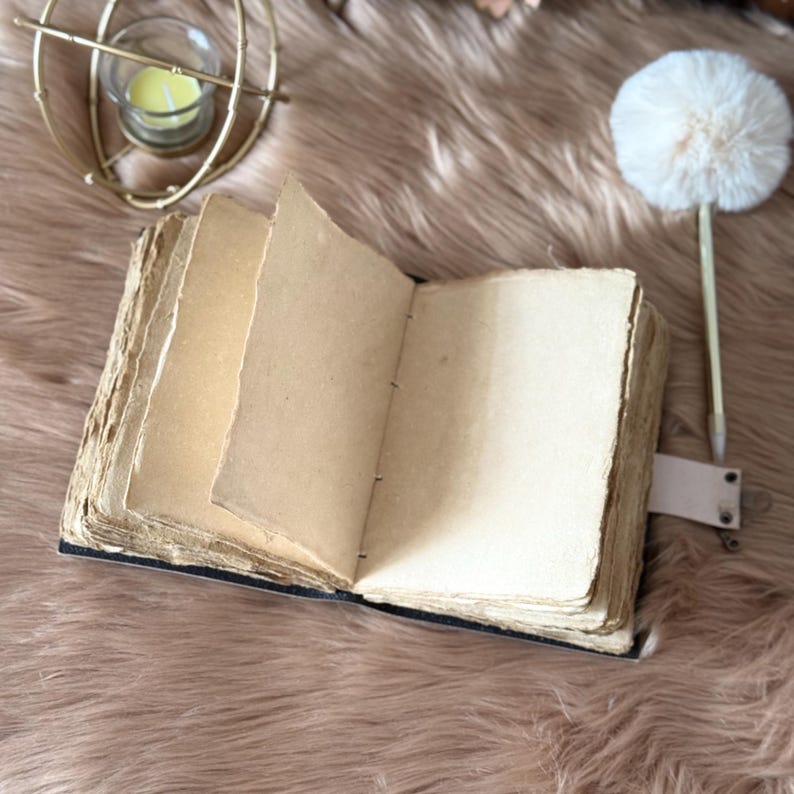 May include: An open, antique-style journal with aged, textured pages. The journal has a dark blue cover and a leather strap closure. A gold candle holder and a fluffy white pen are in the background.