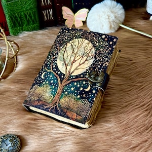 May include: A decorative journal with a celestial design. The cover features a large moon, a tree, and stars against a dark blue background. The journal has a metal clasp and a leather strap. The pages have a vintage, aged look.
