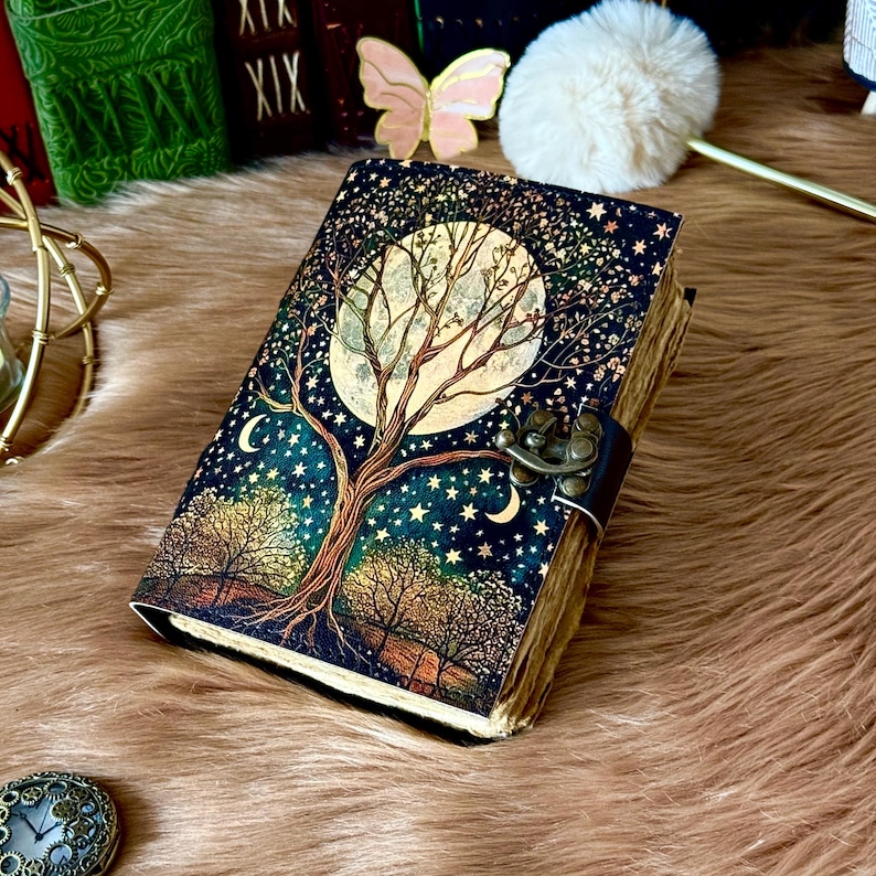 May include: A decorative journal with a celestial design. The cover features a large moon, a tree, and stars against a dark blue background. The journal has a metal clasp and a leather strap. The pages have a vintage, aged look.