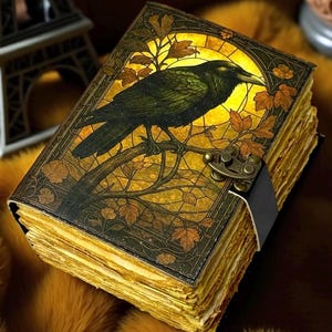 Peut inclure: Un journal de style vintage avec une illustration de corbeau sur la couverture. La couverture présente un motif de vitrail avec un corbeau noir perché sur une branche, sur fond jaune. Le journal a une fermeture à lanière en cuir et du papier vieilli.