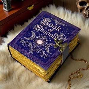 May include: A rectangular journal with a deep purple cover featuring the gold-coloured text Book of Shadows, a pentagram, and crescent moon motifs. The book has a brass-toned metal latch closure and thick, deckled-edge pages with a golden finish.