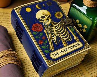 May include: A blue leather journal with a skeleton illustration and the words "THE OVERTHINKER". The cover features a skeleton, roses, and celestial elements. The journal has a metal clasp and stitched binding.