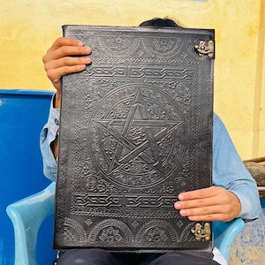 May include: Black leather journal with an embossed pentagram design. The cover features Celtic knotwork and a metal clasp. The journal is held in front of a person, suggesting its size and craftsmanship.