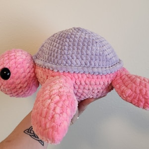 Puede incluir: Un juguete de peluche de tortuga tejido a crochet con un cuerpo rosa y un caparazón morado.