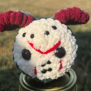 May include: A handmade, cream-colored pom-pom with black eyes and a red smile, resembling a hockey mask. The pom-pom has red and gray accents and sits atop a jar.