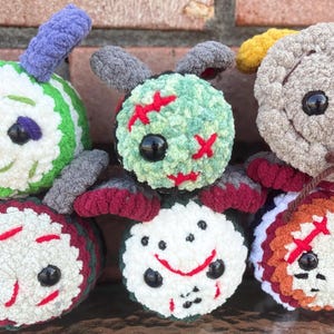 May include: A collection of six handmade, plush, round creatures with stitched details. Each has a unique color scheme, including green, white, red, and orange, with black button eyes and antenna-like appendages. The creatures have a whimsical, cartoonish appearance.