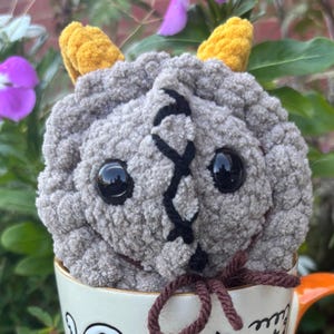 May include: A handmade, gray crocheted creature with black stitched details and large black eyes. It has yellow horns and is sitting in a white mug with an orange handle. A brown yarn bow is tied at the bottom.