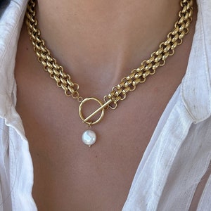 Könnte beinhalten: Eine goldene Kettenkette mit einem großen, runden, weißen Perlenanhänger. Die Kette hat einen Toggle-Verschluss.