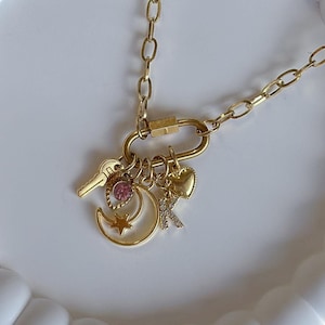 Op de afbeelding: Een gouden ketting met een bedel in de vorm van een halve maan, een ster, een sleutel, een hart en een roze edelsteen. De ketting ligt op een witte achtergrond.