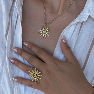 Könnte beinhalten: Eine Person trägt ein weißes Hemd mit einer goldenen Sonnenanhängerkette und einem goldenen Sonnenring. Die Person trägt auch ein Perlenarmband mit Charms.