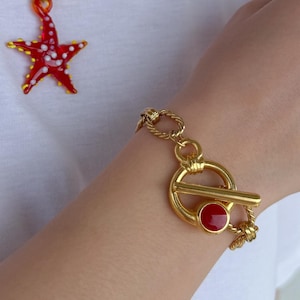 Op de afbeelding: Een gouden ketting met een rode zeester-hanger en een bijpassende gouden armband met een rode ronde hanger. Beide stukken hebben een toggle-sluiting.