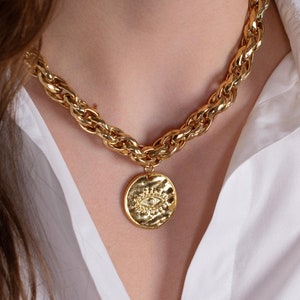 Puede incluir: Un collar de cadena dorada con un colgante grande y redondo con un diseño de ojo texturizado.