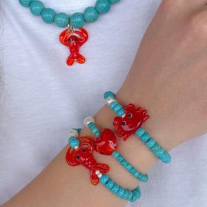 Puede incluir: Un collar de cuentas turquesa con un dije de langosta, y una pulsera de cuentas turquesa con un dije de langosta. La pulsera está superpuesta con otras dos pulseras de cuentas turquesa.