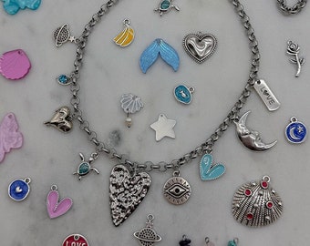 May include: A silver chain necklace with a variety of charms, including hearts, stars, butterflies, seashells, and a small eye. The charms are silver, blue, pink, red, and yellow. The necklace is on a white marble surface.